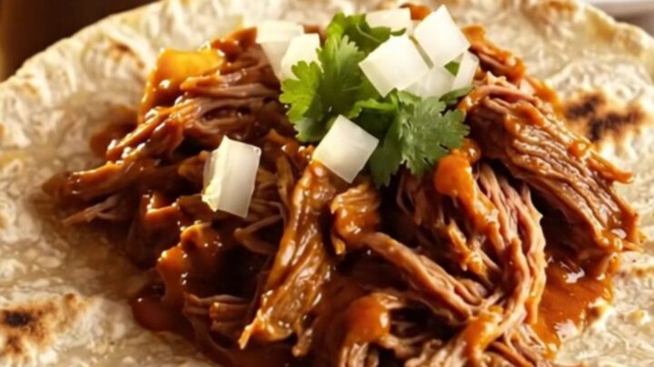 Close up of shredded Chile Colorado beef in a rich red sauce inside a rustic clay-brown slow cooker.
