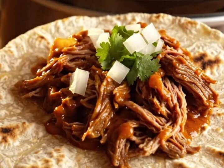 Close up of shredded Chile Colorado beef in a rich red sauce inside a rustic clay-brown slow cooker.