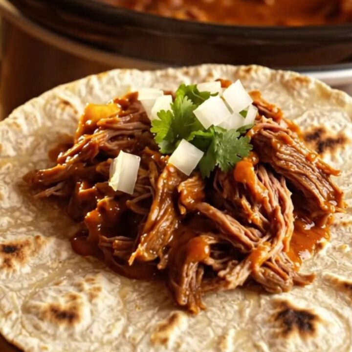Close up of shredded Chile Colorado beef in a rich red sauce inside a rustic clay-brown slow cooker.