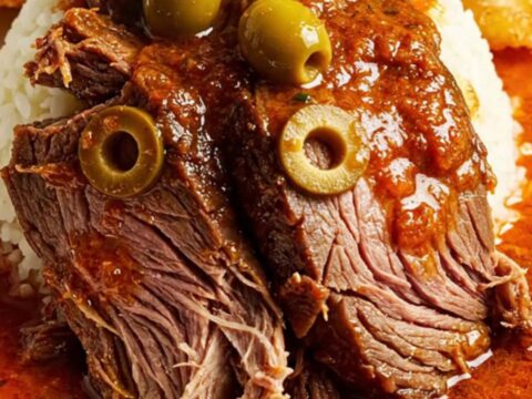 A close-up of a slow-cooked Carne Mechada beef roast sliced on a plate with vibrant green olives visible inside.