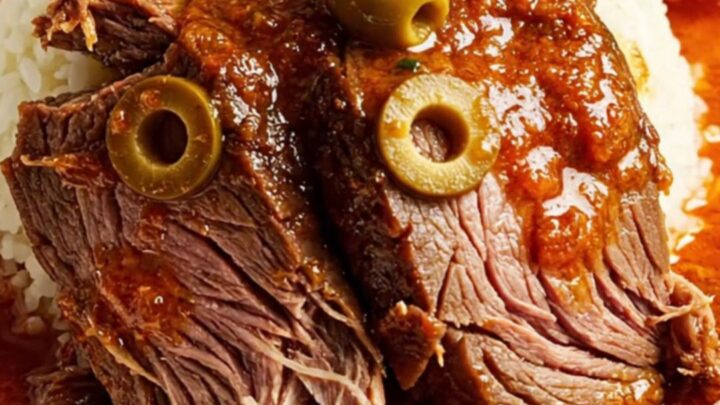 A close-up of a slow-cooked Carne Mechada beef roast sliced on a plate with vibrant green olives visible inside.