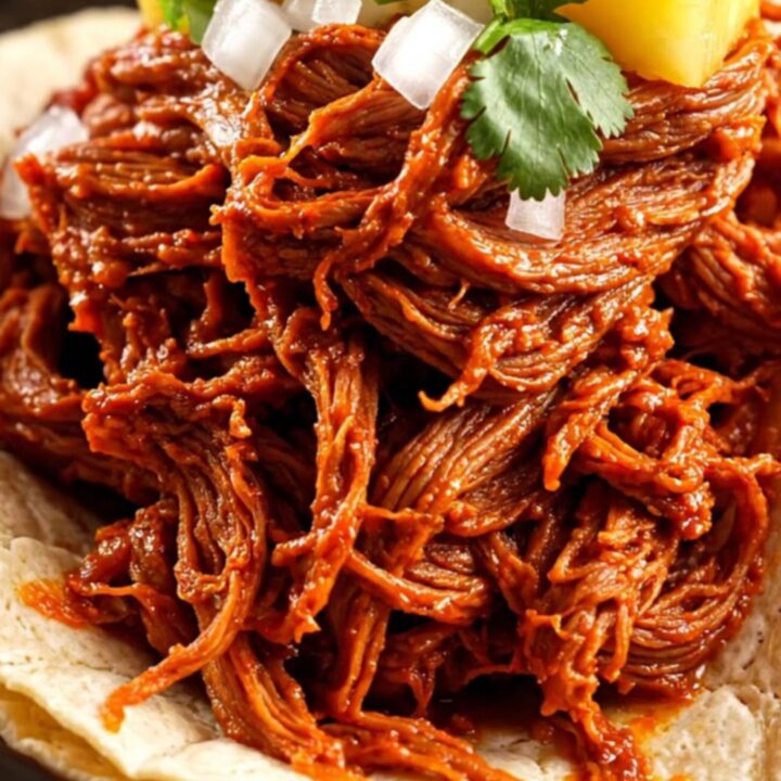 A close-up of shredded red Al Pastor beef tacos topped with pineapple, onion, and cilantro on corn tortillas.