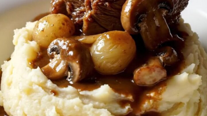 A close-up of tender beef short ribs in a rich, mahogany sauce with pearl onions and mushrooms in a Crockpot.