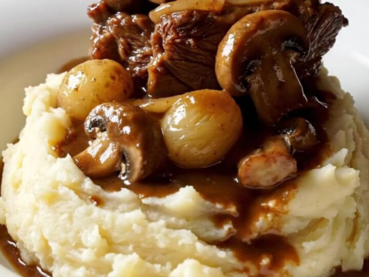 A close-up of tender beef short ribs in a rich, mahogany sauce with pearl onions and mushrooms in a Crockpot.