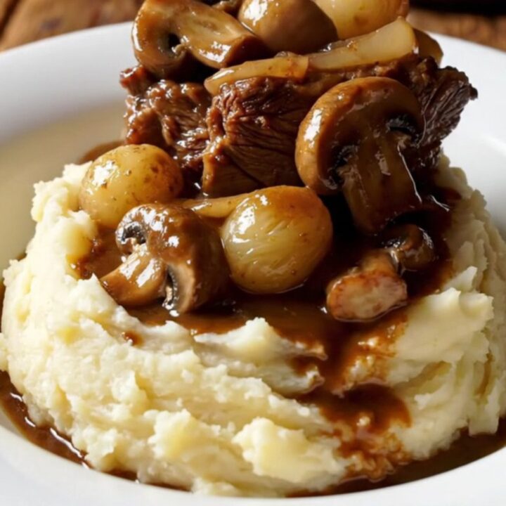 A close-up of tender beef short ribs in a rich, mahogany sauce with pearl onions and mushrooms in a Crockpot.