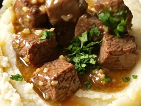 A close-up shot of fork-tender steak bites in a glossy garlic butter sauce inside a black ceramic slow cooker.