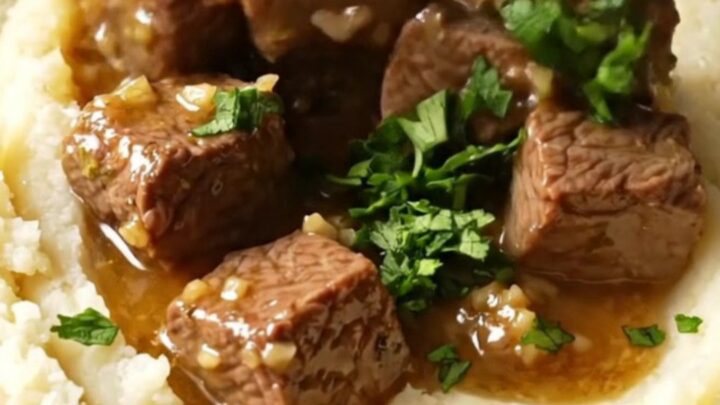 A close-up shot of fork-tender steak bites in a glossy garlic butter sauce inside a black ceramic slow cooker.