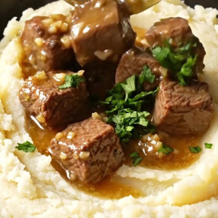 A close-up shot of fork-tender steak bites in a glossy garlic butter sauce inside a black ceramic slow cooker.
