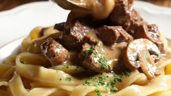 A close-up of creamy ribeye Stroganoff served over buttered egg noodles in a cream stoneware bowl.
