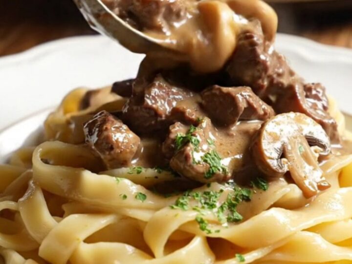 A close-up of creamy ribeye Stroganoff served over buttered egg noodles in a cream stoneware bowl.