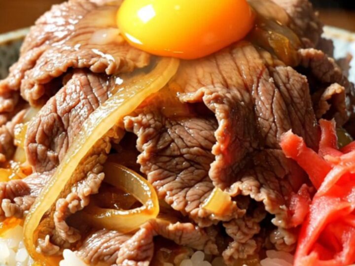 A close-up of a steaming bowl of Japanese Gyudon with tender beef, onions, and a vibrant egg yolk.