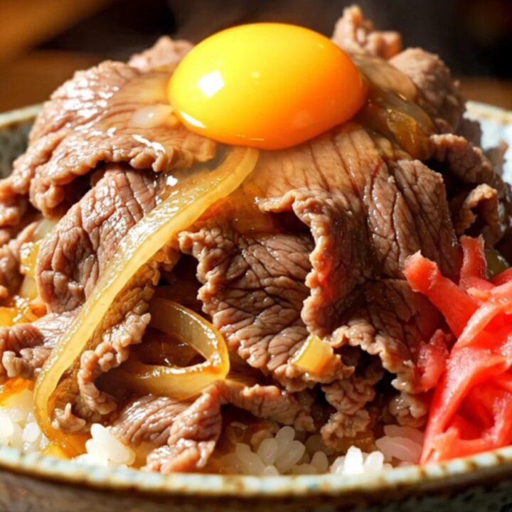 A close-up of a steaming bowl of Japanese Gyudon with tender beef, onions, and a vibrant egg yolk.