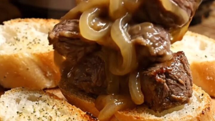 A close-up of tender steak bites in a black ceramic slow cooker covered in melted, bubbly Gruyère cheese.