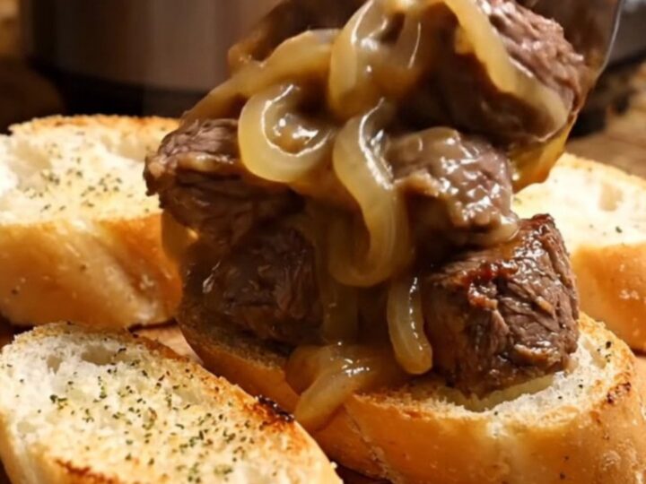 A close-up of tender steak bites in a black ceramic slow cooker covered in melted, bubbly Gruyère cheese.