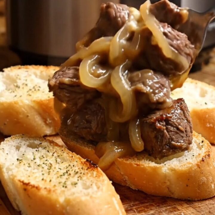 A close-up of tender steak bites in a black ceramic slow cooker covered in melted, bubbly Gruyère cheese.