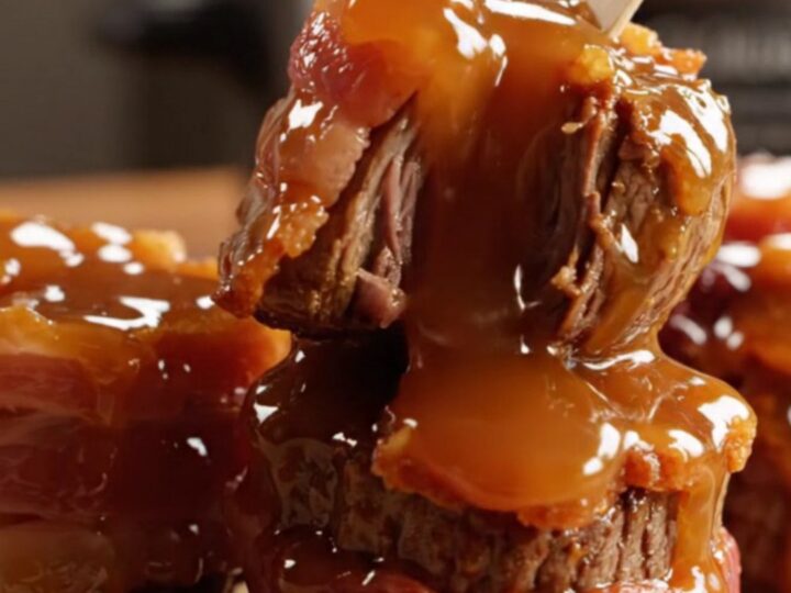 A close-up of bacon-wrapped steak bites glazed in a rich, shiny bourbon sauce on a dark platter.