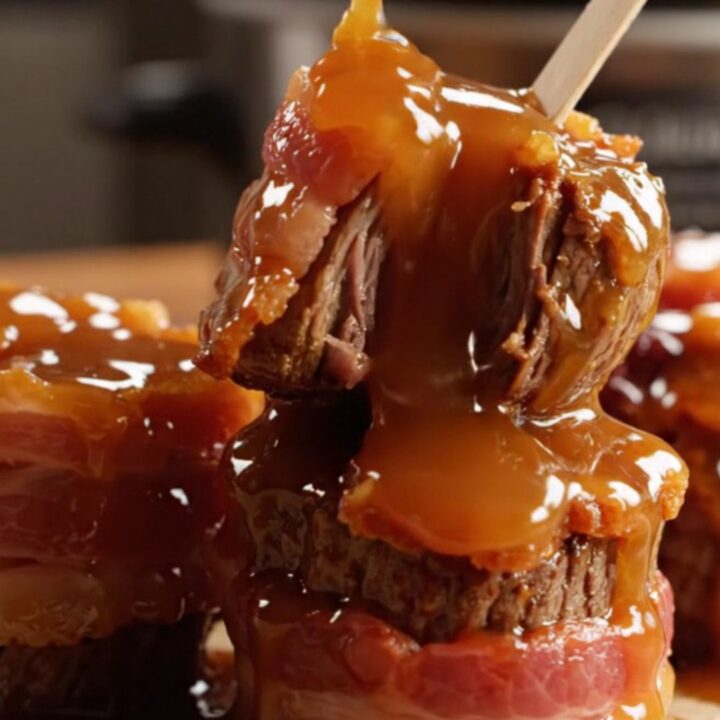 A close-up of bacon-wrapped steak bites glazed in a rich, shiny bourbon sauce on a dark platter.
