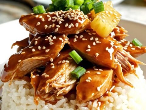 Close-up of shredded pineapple teriyaki chicken served over rice in a bowl, garnished with green onions and sesame seeds.