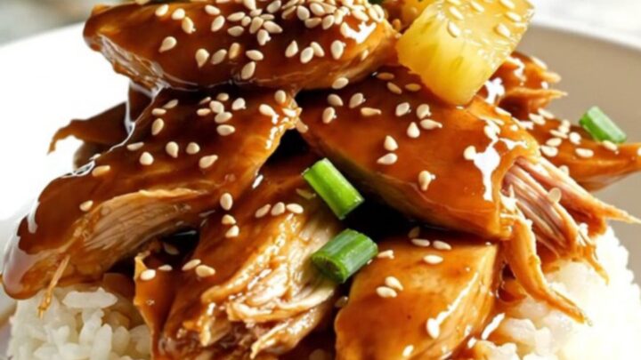 Close-up of shredded pineapple teriyaki chicken served over rice in a bowl, garnished with green onions and sesame seeds.