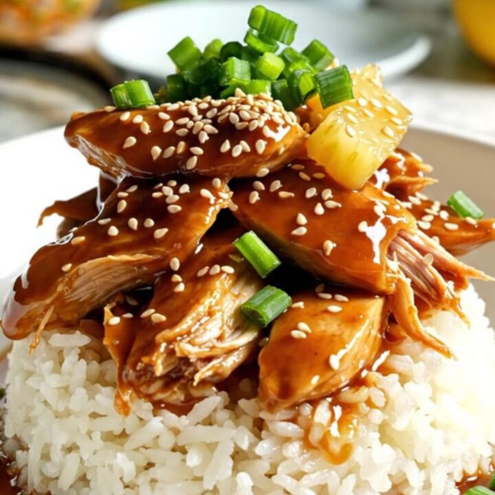 Close-up of shredded pineapple teriyaki chicken served over rice in a bowl, garnished with green onions and sesame seeds.