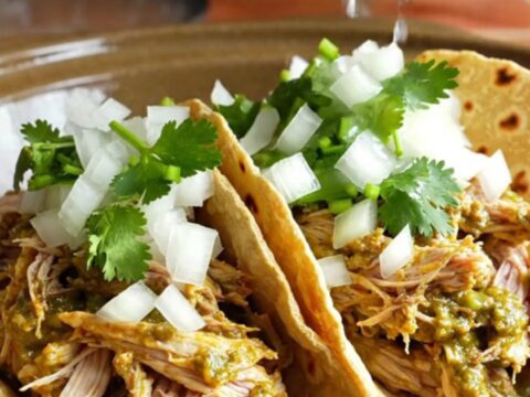 A close-up of shredded chicken in a creamy green salsa verde sauce served in warm corn tortillas with cilantro.