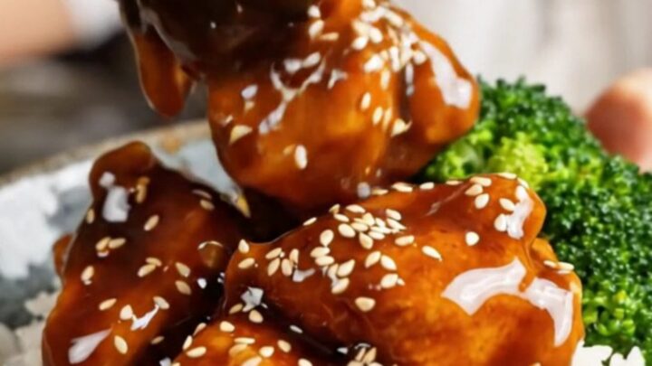 A steaming, glossy bowl of General Tso's chicken with broccoli and sesame seeds over white rice.