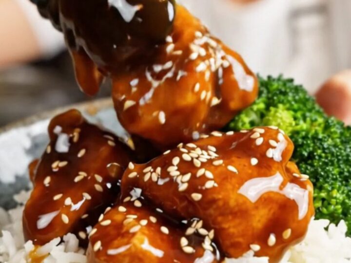 A steaming, glossy bowl of General Tso's chicken with broccoli and sesame seeds over white rice.