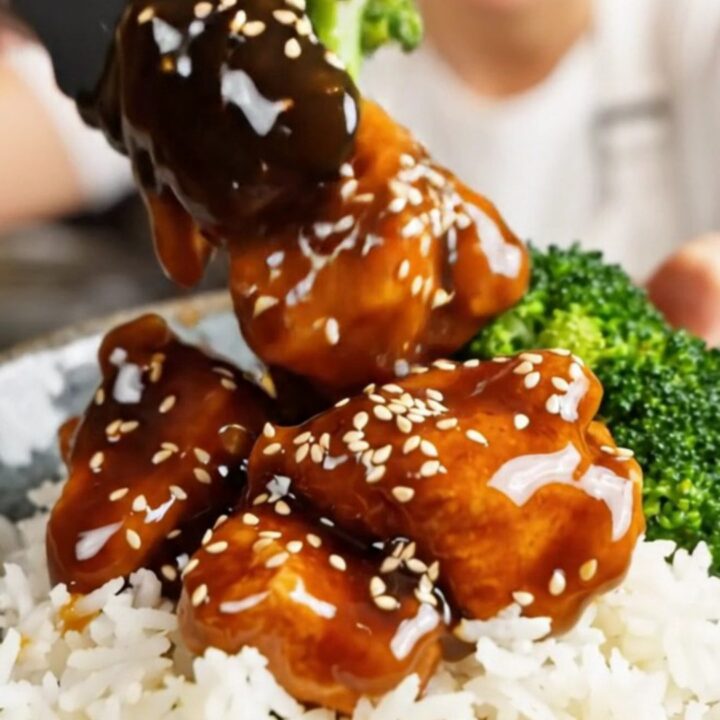 A steaming, glossy bowl of General Tso's chicken with broccoli and sesame seeds over white rice.