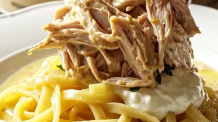 A close-up of tender shredded chicken in a creamy Dijon sauce with fresh tarragon in a cream-colored stoneware slow cooker.