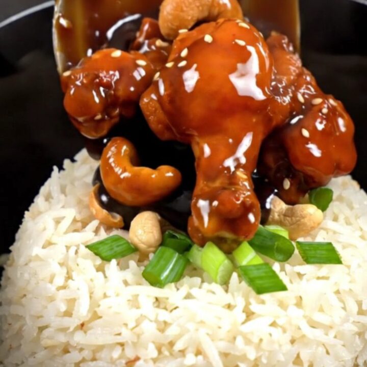 A close-up of glossy Kung Pao chicken with roasted cashews and scallions served in a bowl.