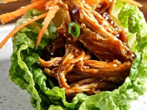A close-up of a lettuce wrap filled with shredded, glossy hoisin chicken, topped with julienned carrots and scallions.