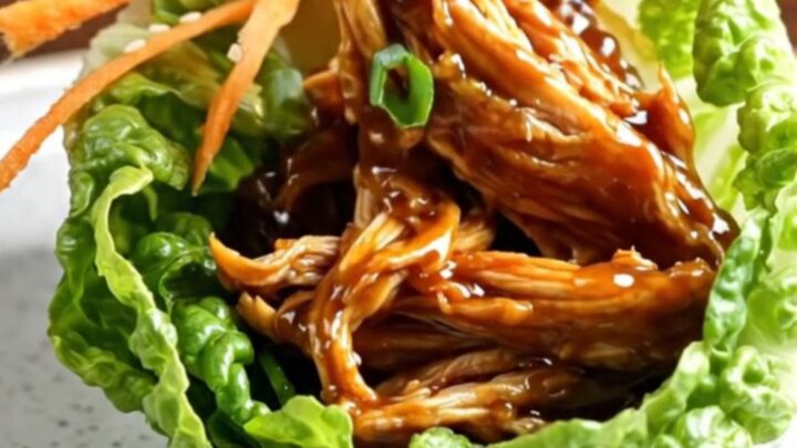 A close-up of a lettuce wrap filled with shredded, glossy hoisin chicken, topped with julienned carrots and scallions.
