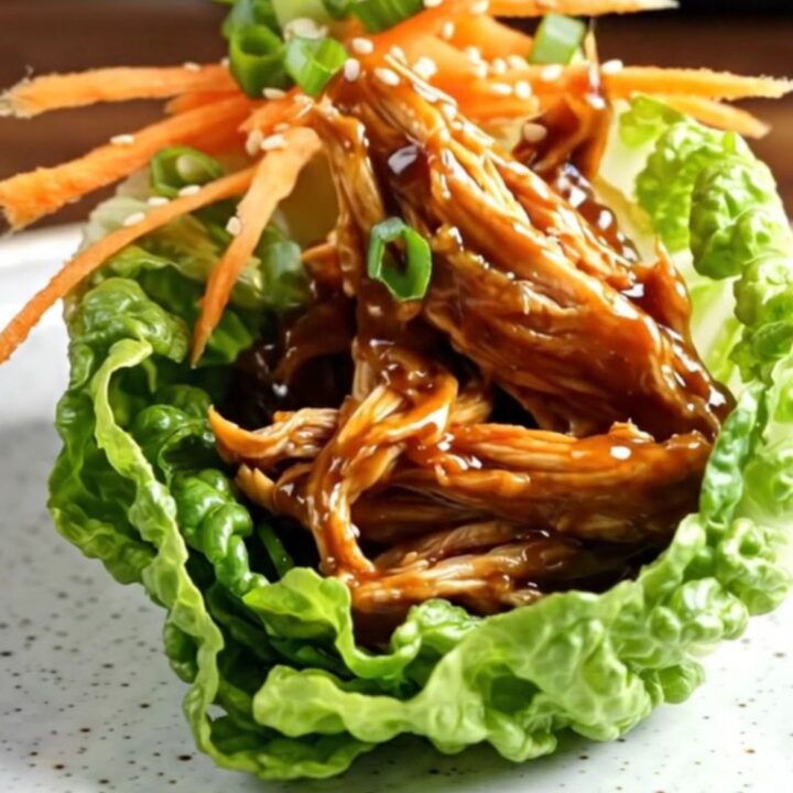 A close-up of a lettuce wrap filled with shredded, glossy hoisin chicken, topped with julienned carrots and scallions.