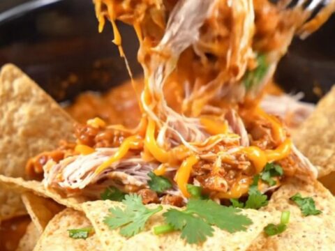A warm red slow cooker filled with creamy shredded chicken, chorizo, and melted white queso, topped with cilantro.