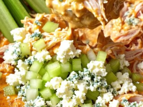 A close-up view of creamy buffalo chicken in a slow cooker, being shredded with two forks, topped with blue cheese crumbles.