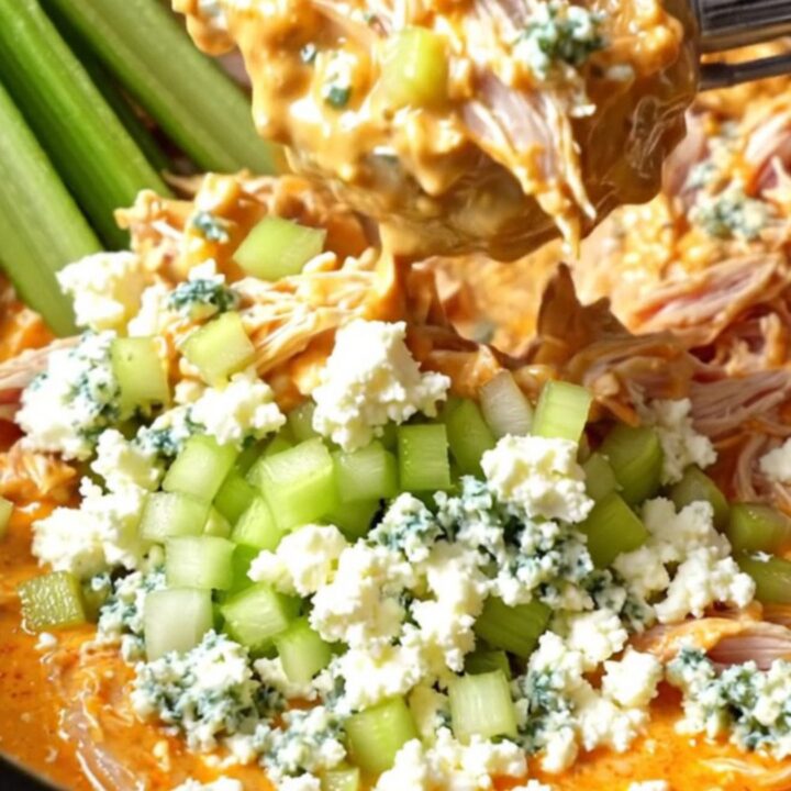 A close-up view of creamy buffalo chicken in a slow cooker, being shredded with two forks, topped with blue cheese crumbles.