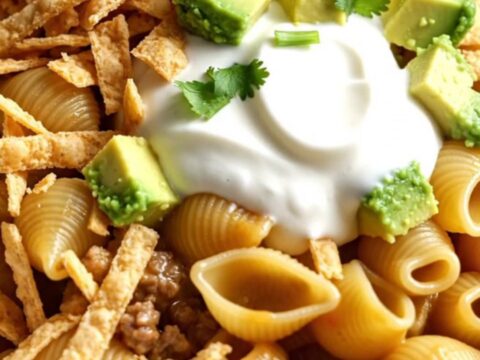 A close-up of cheesy taco pasta shells in a ceramic crockpot topped with avocado, cilantro, and tortilla strips.