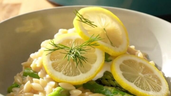 A wide ceramic bowl filled with creamy lemon orzo, tender chicken, and bright green asparagus spears.