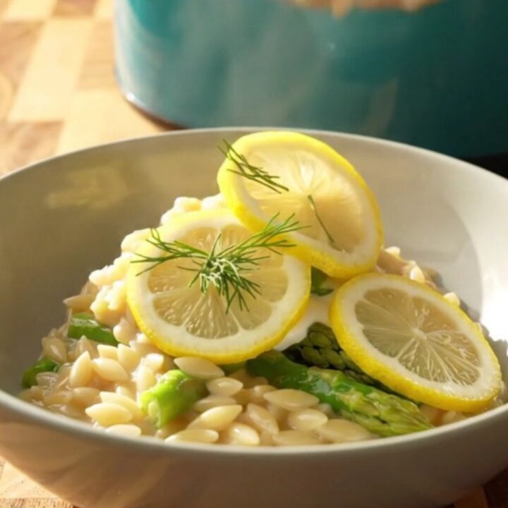 A wide ceramic bowl filled with creamy lemon orzo, tender chicken, and bright green asparagus spears.