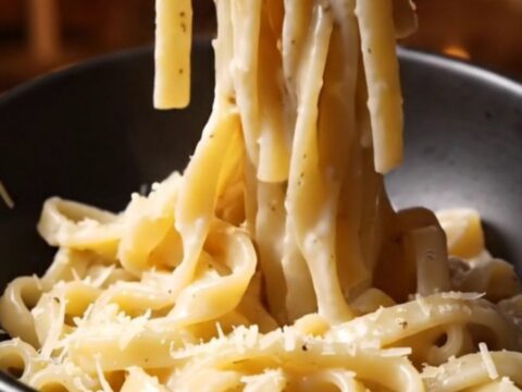 A close-up of glossy bucatini pasta coated in a creamy Pecorino Romano sauce with visible black pepper specks.