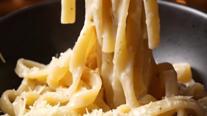 A close-up of glossy bucatini pasta coated in a creamy Pecorino Romano sauce with visible black pepper specks.