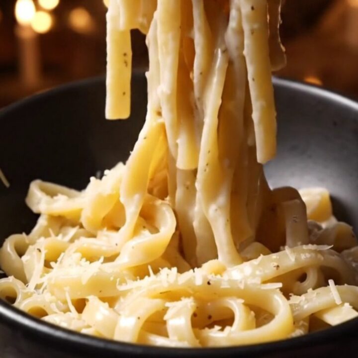 A close-up of glossy bucatini pasta coated in a creamy Pecorino Romano sauce with visible black pepper specks.