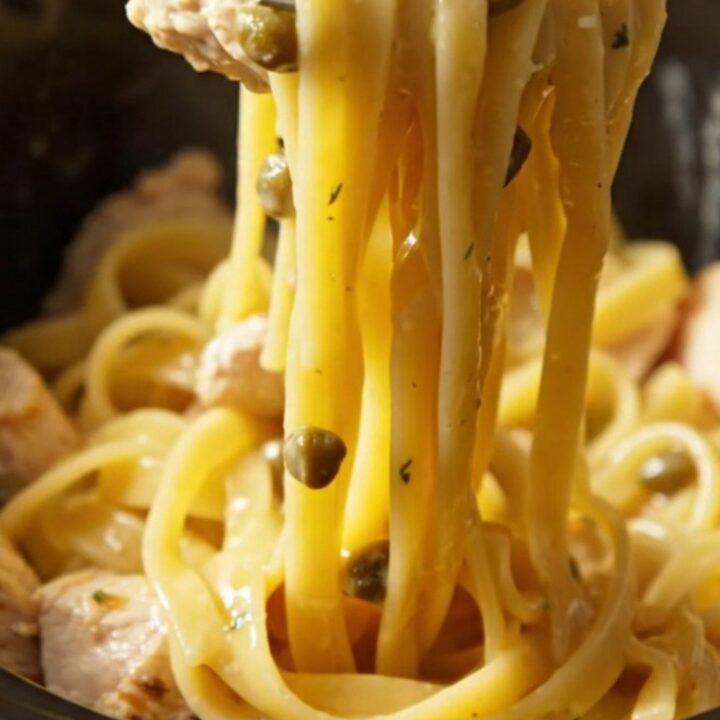 Close-up of creamy chicken piccata pasta with capers and lemon slices in a bright setting.