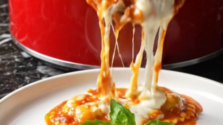 A close-up of a ruby-red crockpot filled with bubbling four-cheese ravioli topped with melted cheese and fresh herbs.
