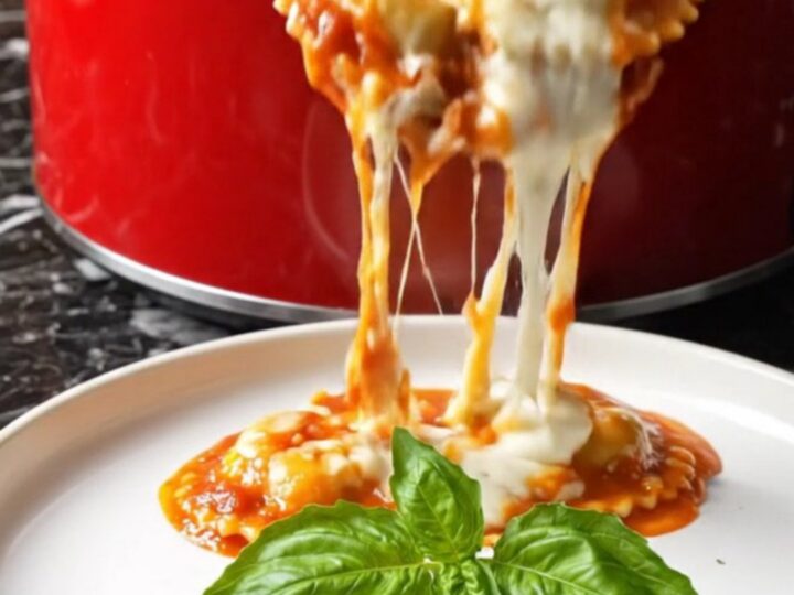 A close-up of a ruby-red crockpot filled with bubbling four-cheese ravioli topped with melted cheese and fresh herbs.