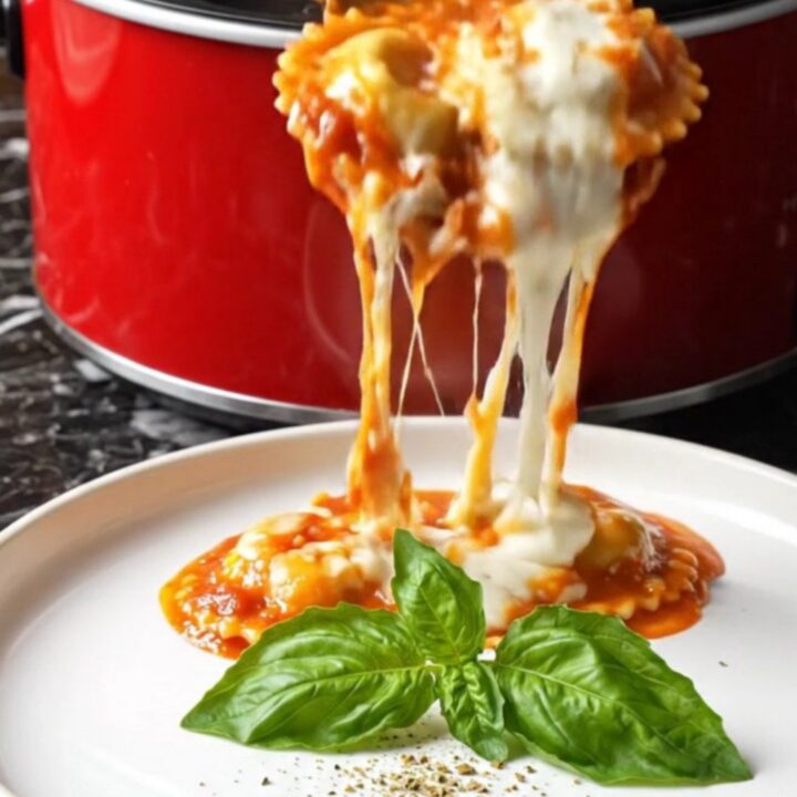 A close-up of a ruby-red crockpot filled with bubbling four-cheese ravioli topped with melted cheese and fresh herbs.