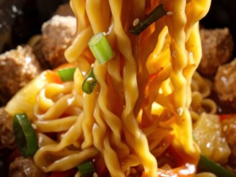 A close-up of a slow cooker filled with glazed pork meatballs, colorful bell peppers, and lo mein noodles.
