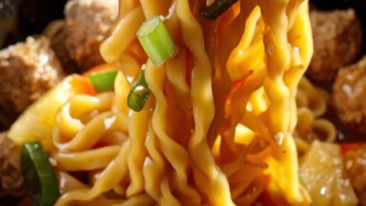 A close-up of a slow cooker filled with glazed pork meatballs, colorful bell peppers, and lo mein noodles.