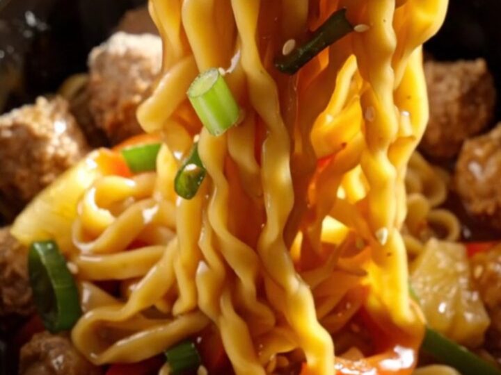 A close-up of a slow cooker filled with glazed pork meatballs, colorful bell peppers, and lo mein noodles.