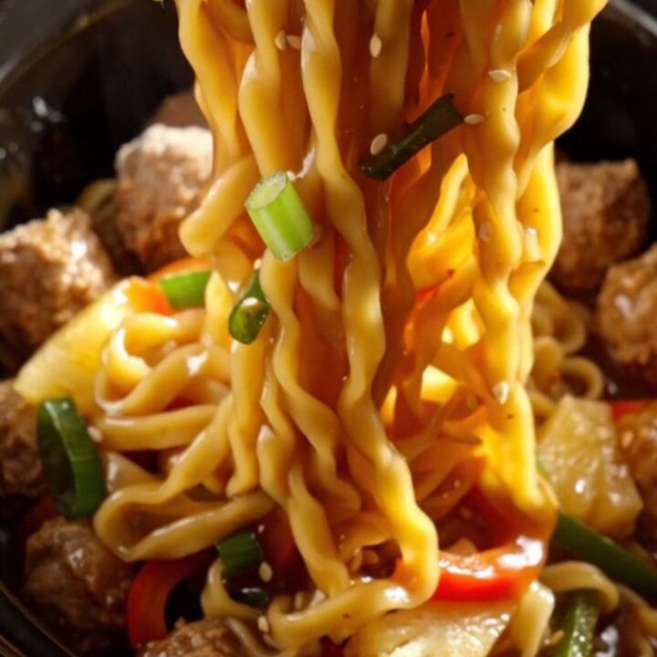 A close-up of a slow cooker filled with glazed pork meatballs, colorful bell peppers, and lo mein noodles.